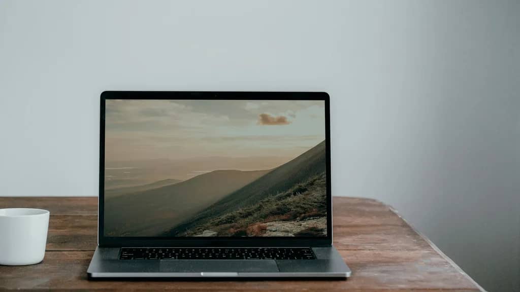 Aufgeklappter Laptop mit Berglandschaft auf dem Bildschirm auf einem Holztisch vor heller Wand, daneben eine weisse Tasse