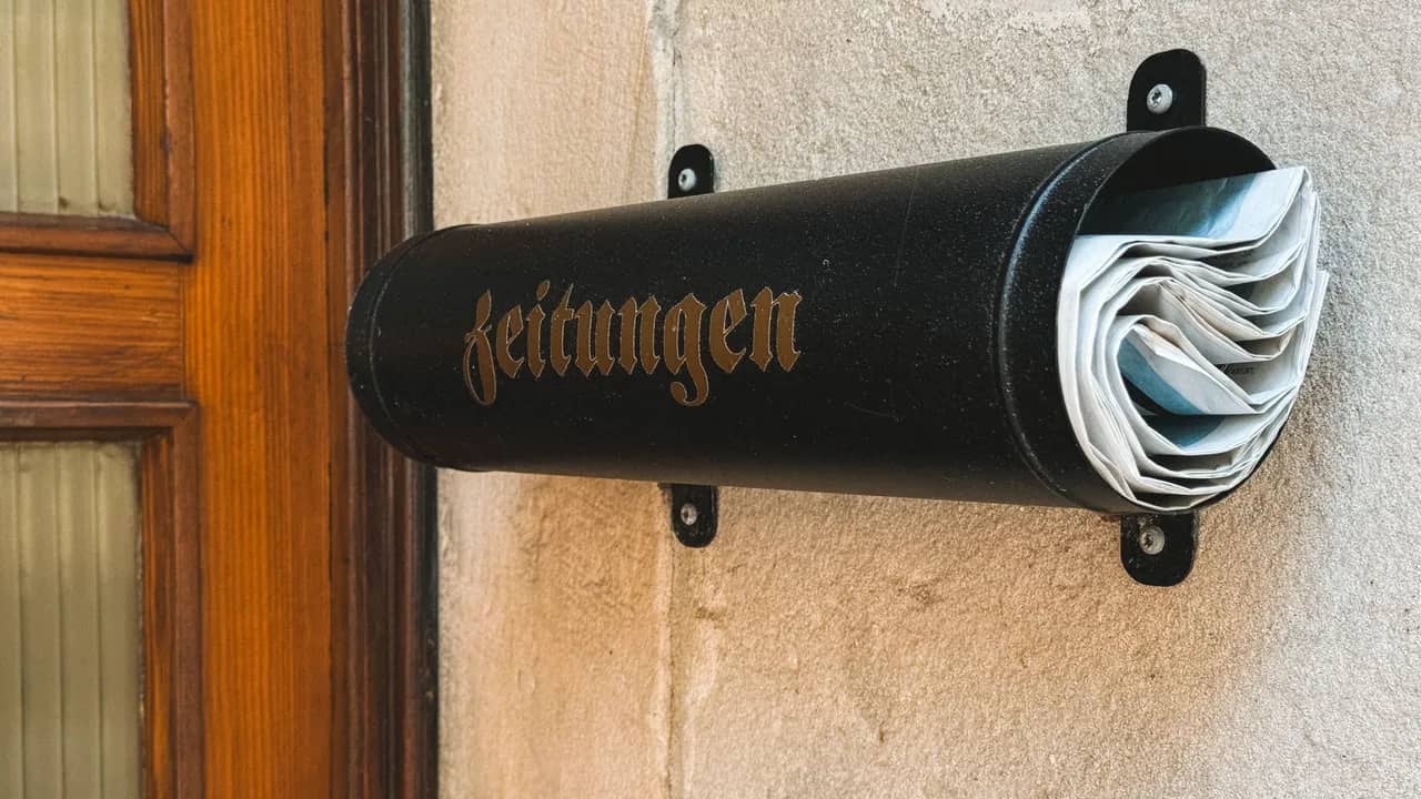 Schwarze Zeitungsrolle mit der Aufschrift «Zeitungen» an einer hellen Hauswand neben einer Holztür, darin gerollte Zeitungen.