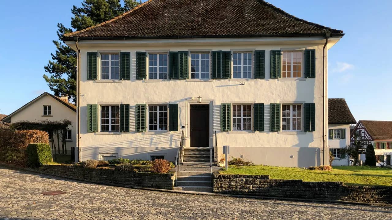 Zweigeschossiges, weiss verputztes Wohnhaus mit grünen Fensterläden und Walmdach, davor eine Aussentreppe, Rasen und Kopfsteinpflaster.